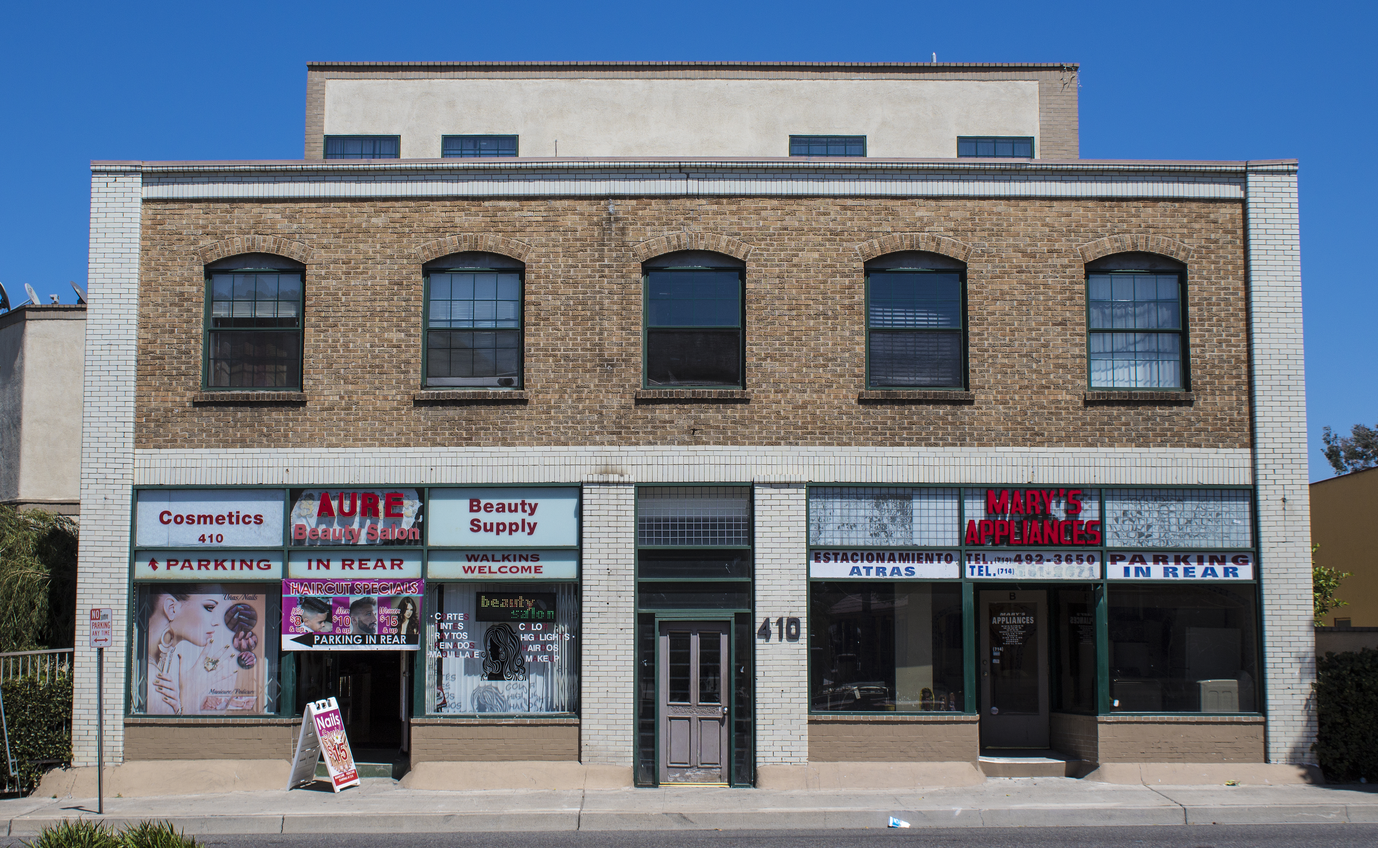 A colored photo of the De Luxe Hotel in 2017 with two storefronts: AURE Beauty Salon and Mary&#039;s Appliances.