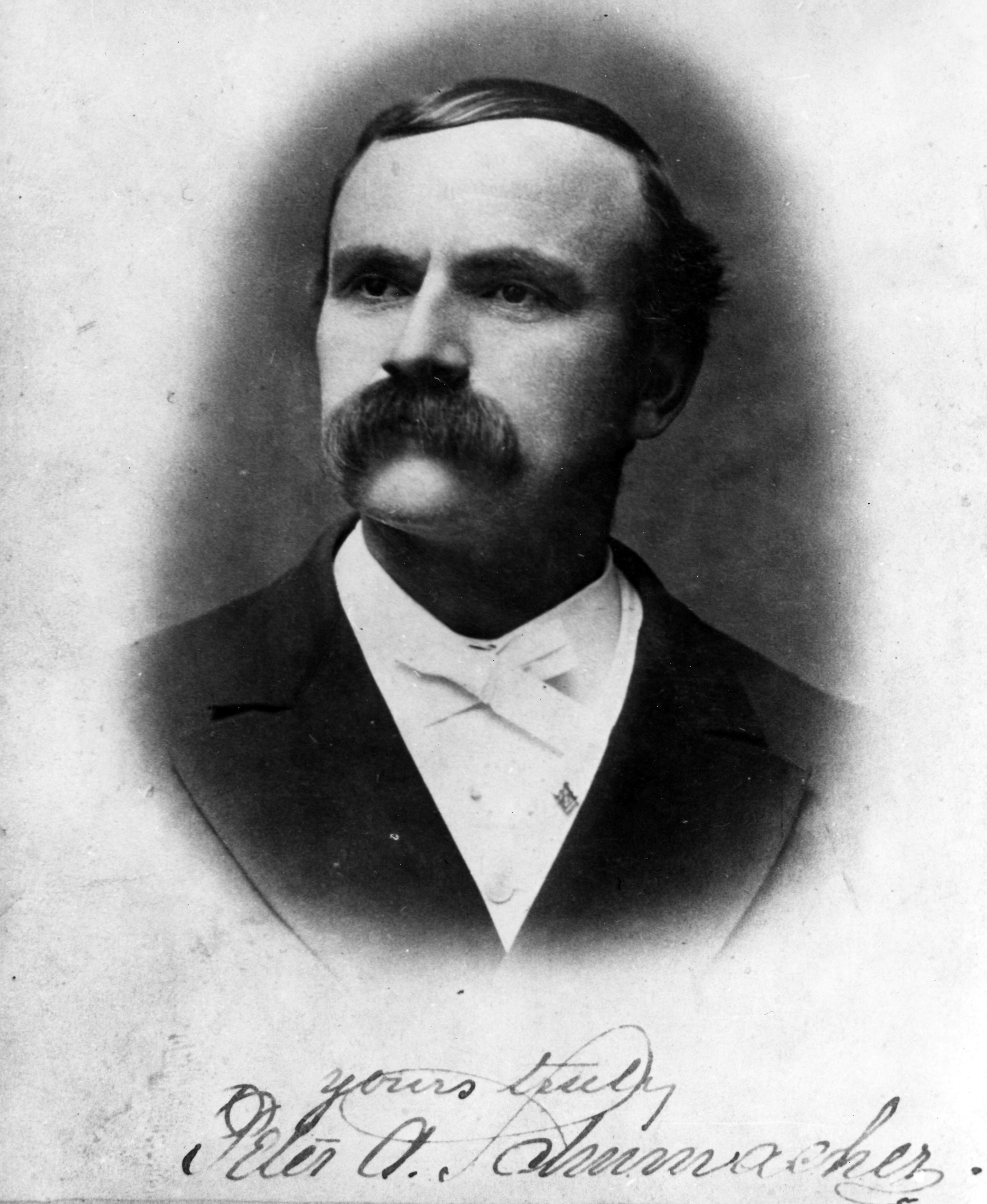A black and white portrait photograph of Peter Schumacher. The bottom of the photo is signed: Yours truly. Peter A. Schumacher.