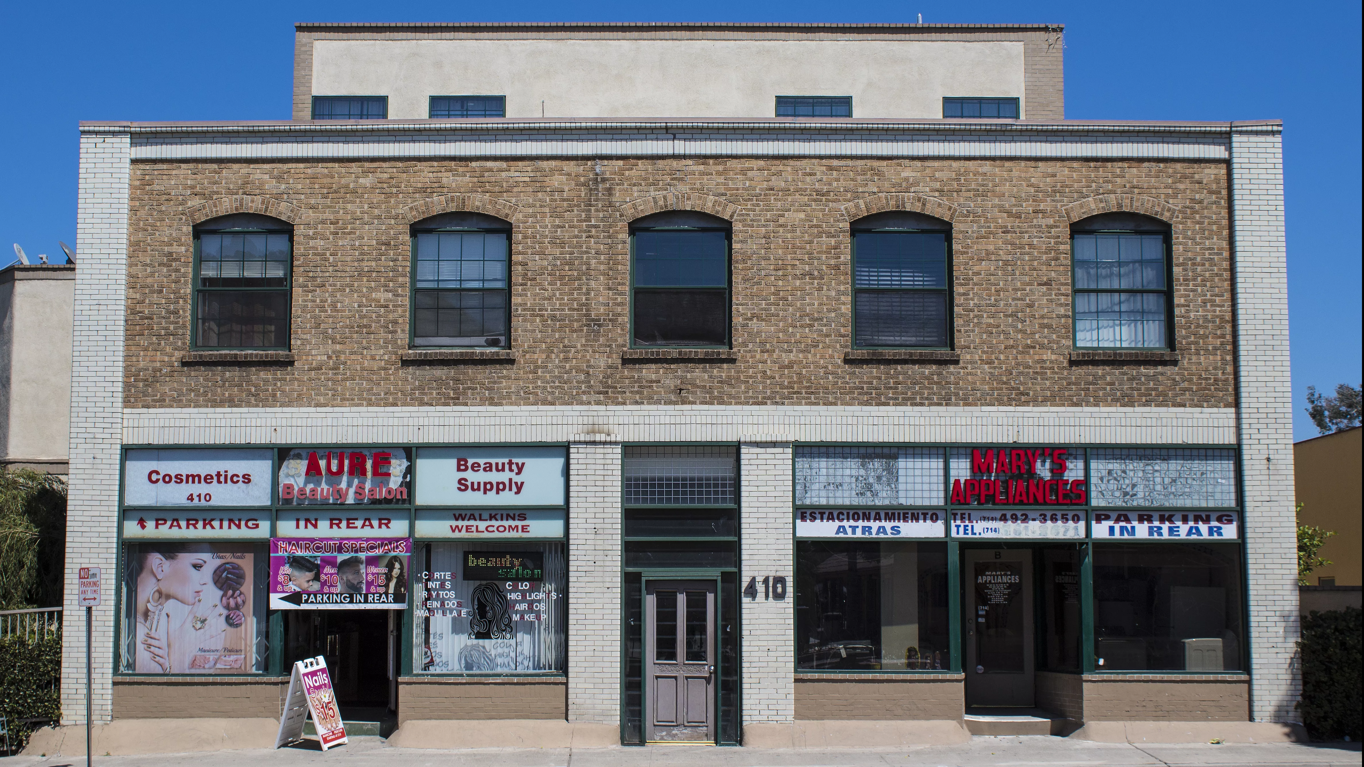 A colored photo of the De Luxe Hotel in 2017 with two storefronts: AURE Beauty Salon and Mary's Appliances.