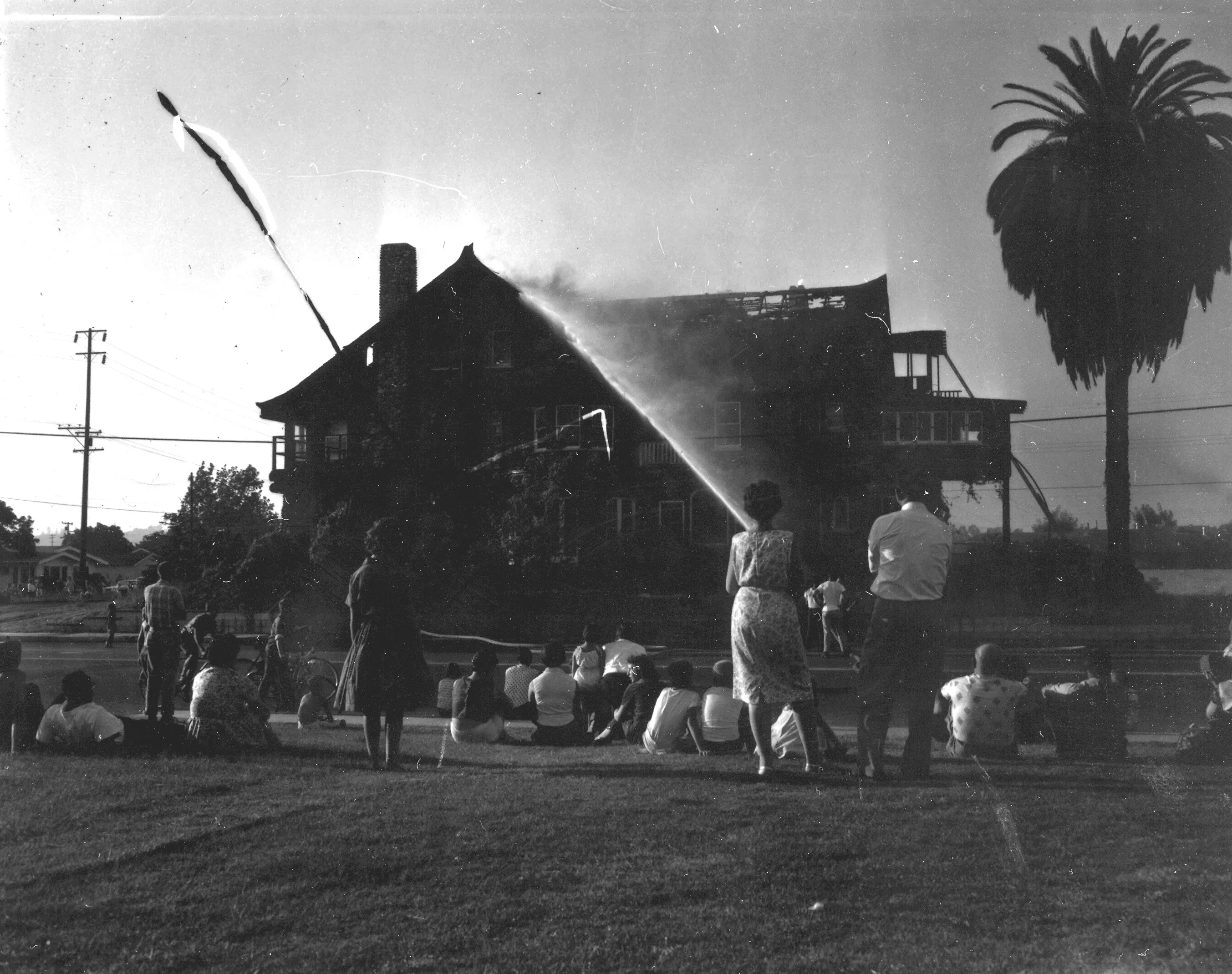 A black and white photograph of the neighborhood families standing and sitting outside watching the Chapman House on fire in 1960.